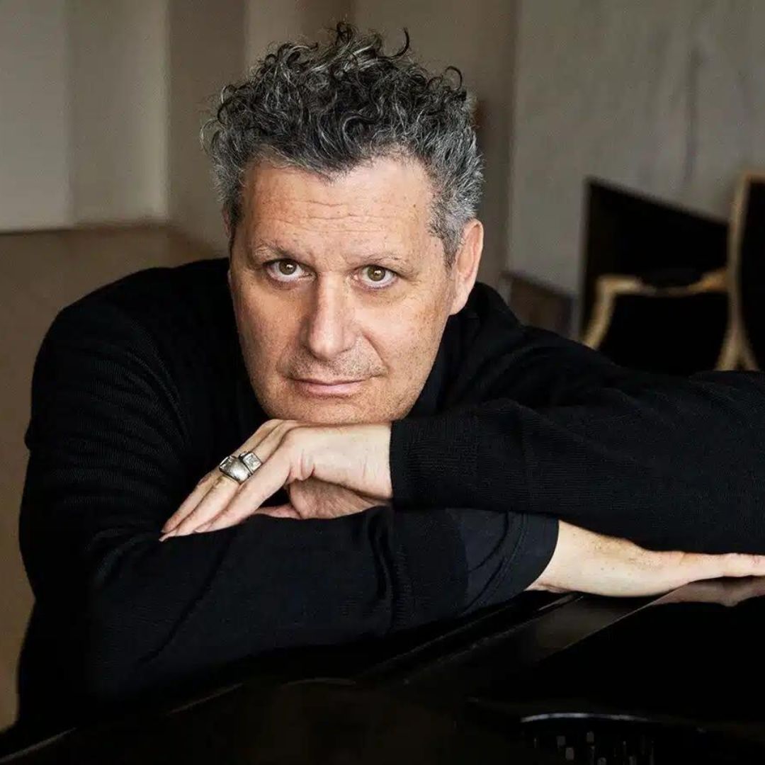 a black and white photo of Isaac Mizrahi, leaning his head on his hand and looking into the camera