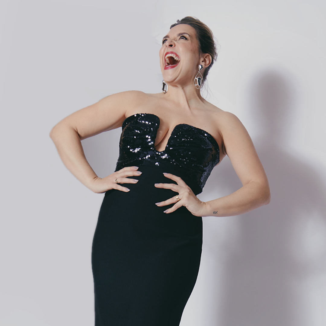 Performer Jessica Vosk in a ballgown laughs on stage with her hands on her hips